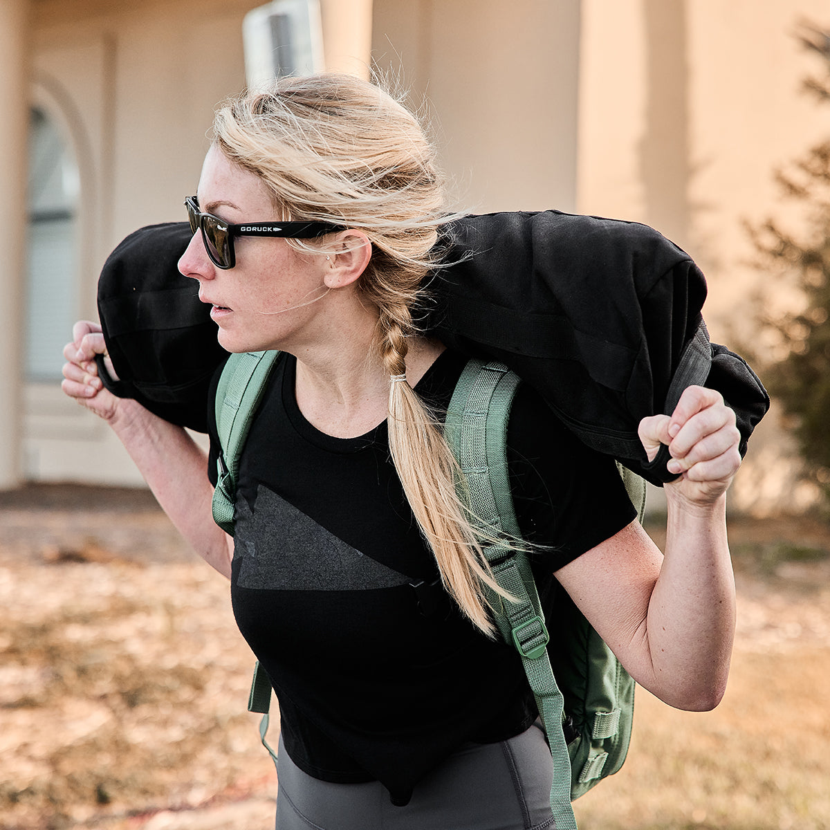 A badass babe with sunglasses strides outdoors, rocking a Women's Cropped Tee - Poly-Blend and a green backpack while confidently carrying a hefty sandbag.