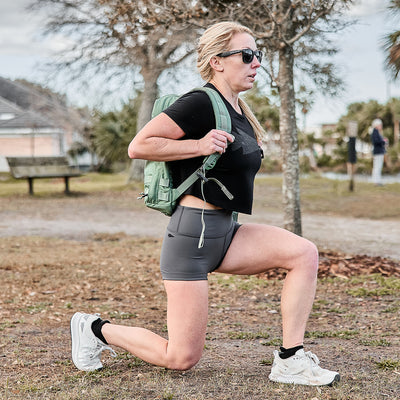 Wearing a Women's Cropped Tee - Poly-Blend, sunglasses, and a backpack, a person in workout attire does lunges outdoors with a badass energy. Surrounded by trees and a bench, the serene backdrop contrasts with the determined vibe of the scene.