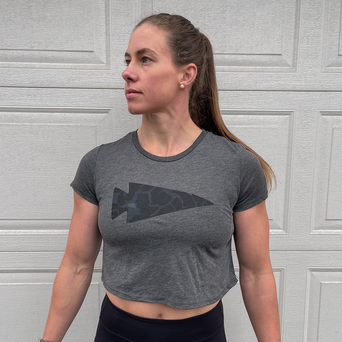 A person wearing a Women's Cropped Tee - Poly-Blend in a super soft gray arrow graphic stands in front of a garage door, looking to the side. Ideal for fierce women ready for their next workout.