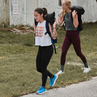 Two women embark on a GORUCK Women's Rough Runner - Electric Blue journey, carrying heavy bags on their shoulders. One wears a sleeveless shirt and blue shoes, while the other dons a long-sleeve shirt and maroon leggings. Both appear focused and determined, treading the grassy path with high mileage resilience.