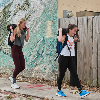 Two women in athletic gear from GORUCK, embodying the Rough Runner spirit, carry sandbags on their shoulders as they walk past a colorful mural on a wooden fence. One is dressed in a gray shirt and burgundy leggings, while the other sports a white top paired with black leggings. Both appear focused and determined. The product featured is the Women's Rough Runner - Electric Blue by GORUCK.