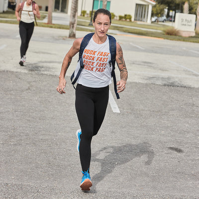 A Women's Rough Runner from GORUCK sprints towards the camera in a parking lot, wearing black leggings and a white sleeveless shirt with text. Sporting Electric Blue athletic shoes and a backpack, they exude energy. Another person looms in the background, while buildings and trees create a gradient density in the distance.