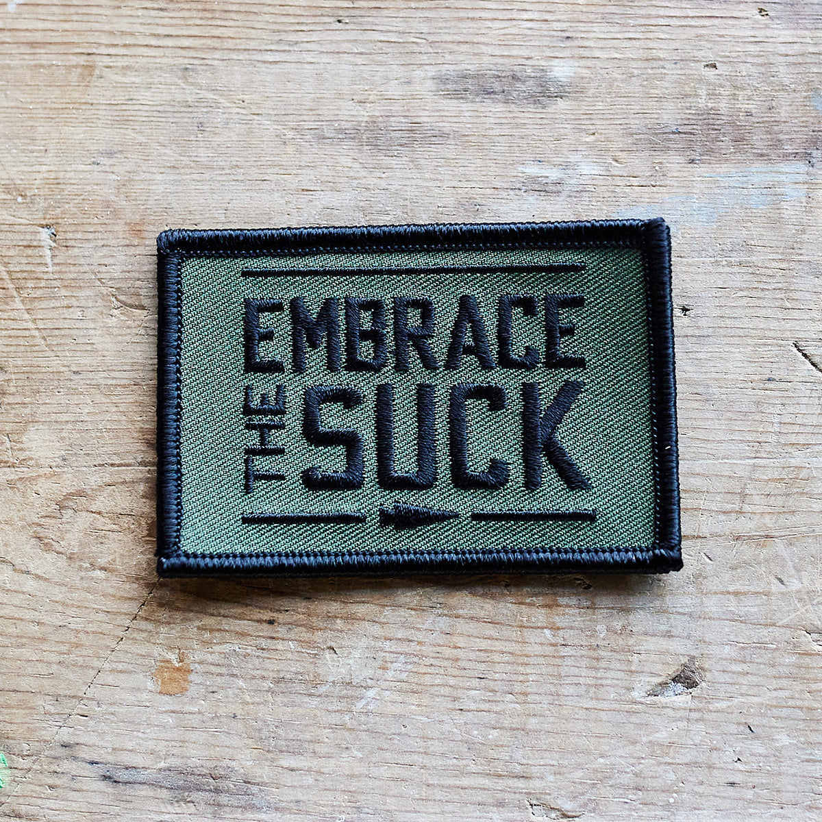 An embroidered patch from vendor-unknown, titled "Patch - Embrace The Suck," is secured with VELCRO® backing and showcases the phrase "Embrace the Suck" prominently in bold black letters, encircled by a striking black border.