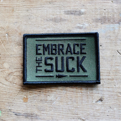 An embroidered patch from vendor-unknown, titled "Patch - Embrace The Suck," is secured with VELCRO® backing and showcases the phrase "Embrace the Suck" prominently in bold black letters, encircled by a striking black border.