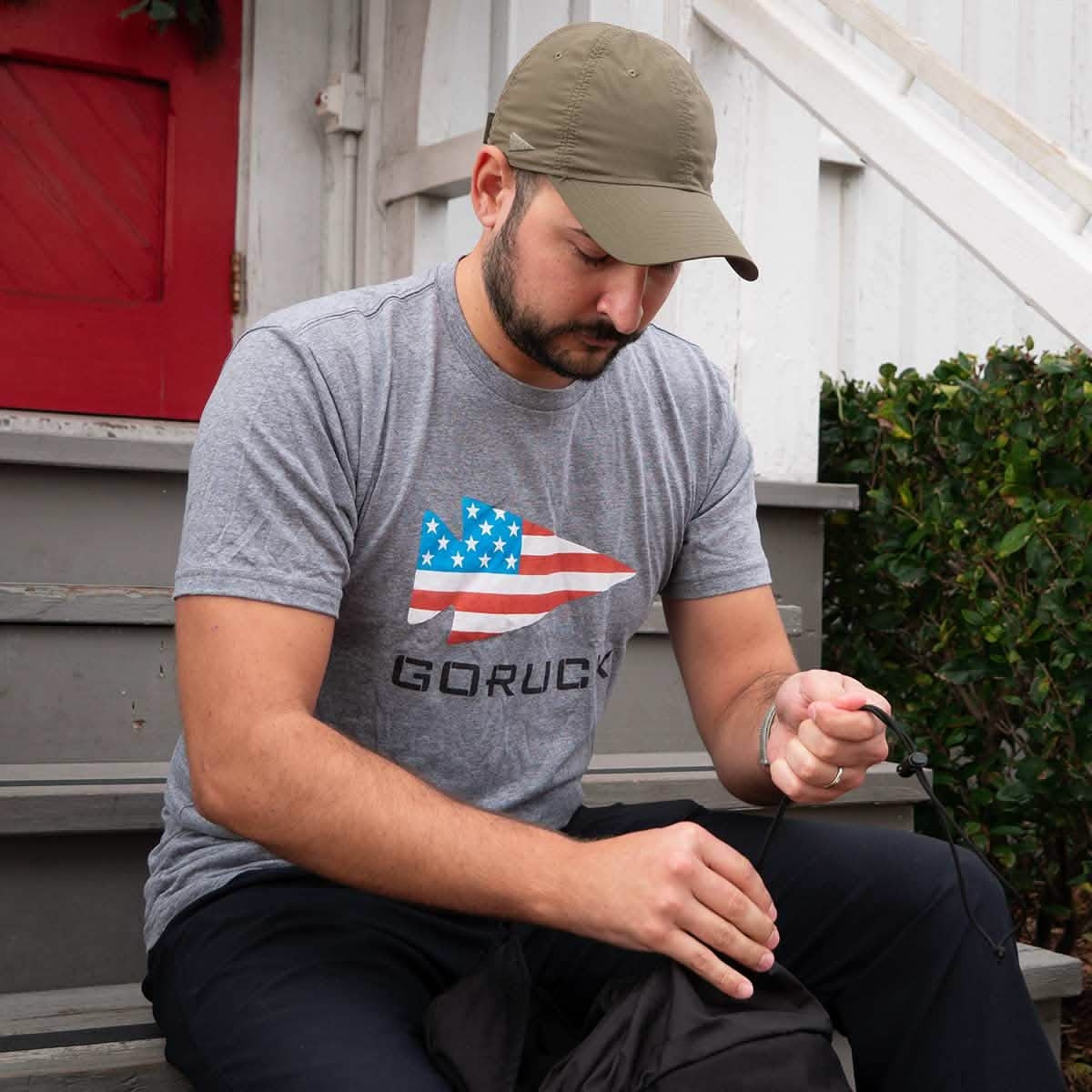 A man in a tri-blend "GORUCK USA" grey T-shirt and cap sits on steps, focused intently on an item in his hands.