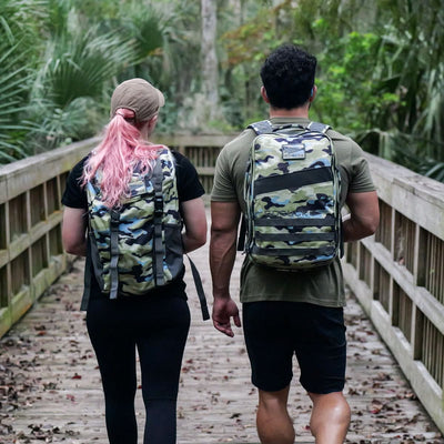 Two individuals with GORUCK Rucker 4.0 camouflage rucksacks stroll across a wooden bridge in a forest. The person on the left sports pink hair, wearing a cap, black shirt, and leggings. Meanwhile, the person on the right has short dark hair and is dressed in a green shirt and black shorts.