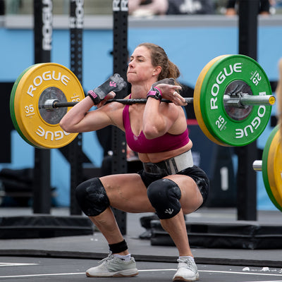 A woman wearing GORUCK's Women's Ballistic Trainers in Lunar Rock and Gum with a Silver Reflective Spearhead, along with athletic gear, performs a front squat with a barbell during a weightlifting competition. The barbell is loaded with yellow and green plates. Intently focused, she is also equipped with knee sleeves, gloves, and a supportive belt.