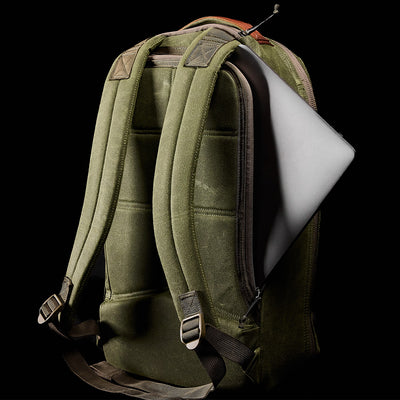 The GORUCK Bullet Ruck - Waxed Canvas in green, featuring padded straps, is displayed against a black background with a laptop partially emerging from its open side pocket.