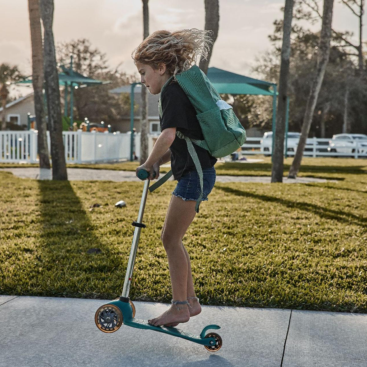 A child rides a scooter on a sunny sidewalk, wearing the durable GORUCK KR1 2.0 - Kid Ruck backpack with reinforced stitching and shorts, amidst the backdrop of trees and houses.