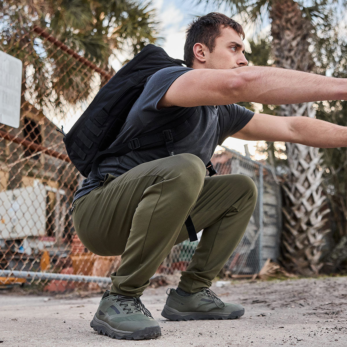 A man outfitted in GORUCK's olive green Men's Performance Joggers, a gray t-shirt, and a black backpack is squatting on a dirt trail. Surrounded by a fence and palm trees, he relishes the comfort of the joggers' moisture-wicking fabric.