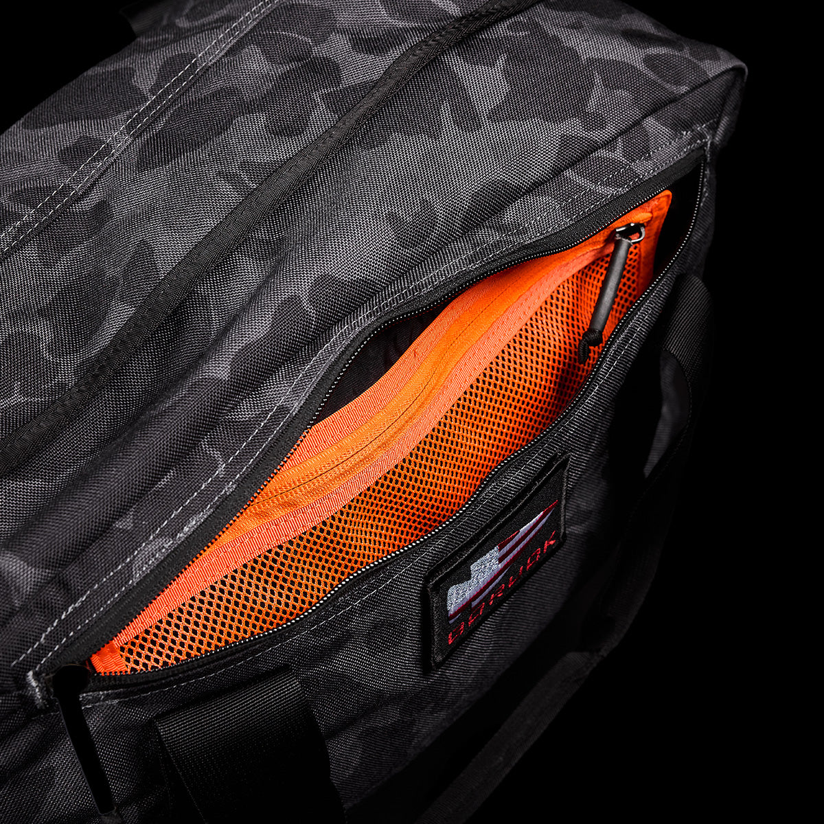 Close-up of a black camouflage-patterned GORUCK Kit Bag with an open pocket revealing a bright orange mesh lining and a small zippered compartment. This durable deployment bag features sturdy handles and includes a shoulder strap, along with a label on the front.