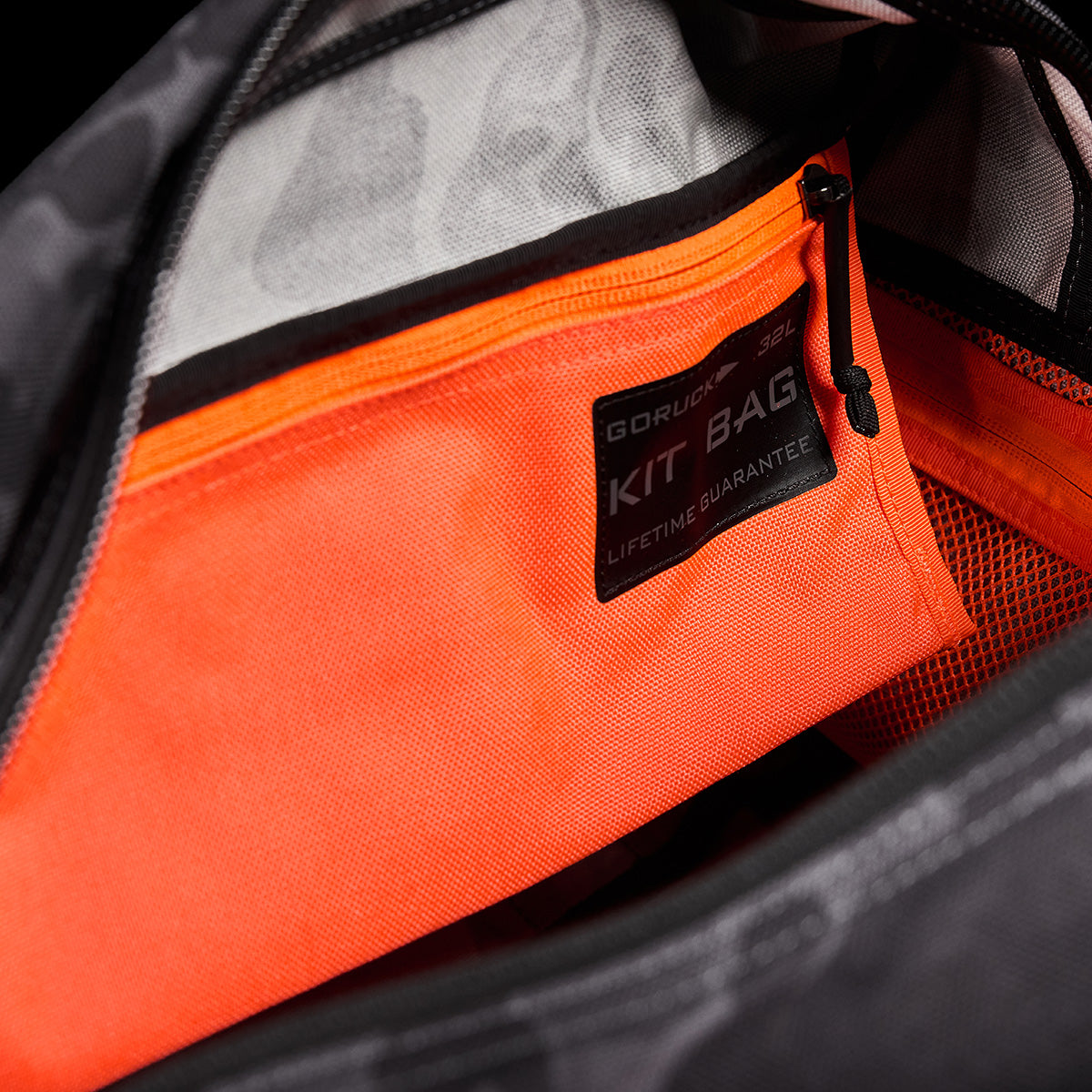 Close-up of an open bag displaying an orange zippered pocket inside. A label on the pocket reads GORUCK Kit Bag: Lifetime Guarantee. The interior of the bag is gray with black accents, and the unzipped zipper reveals a vibrant orange inner pocket, reminiscent of Special Forces Flight Bags.