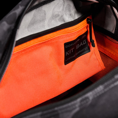 Close-up of an open bag displaying an orange zippered pocket inside. A label on the pocket reads GORUCK Kit Bag: Lifetime Guarantee. The interior of the bag is gray with black accents, and the unzipped zipper reveals a vibrant orange inner pocket, reminiscent of Special Forces Flight Bags.