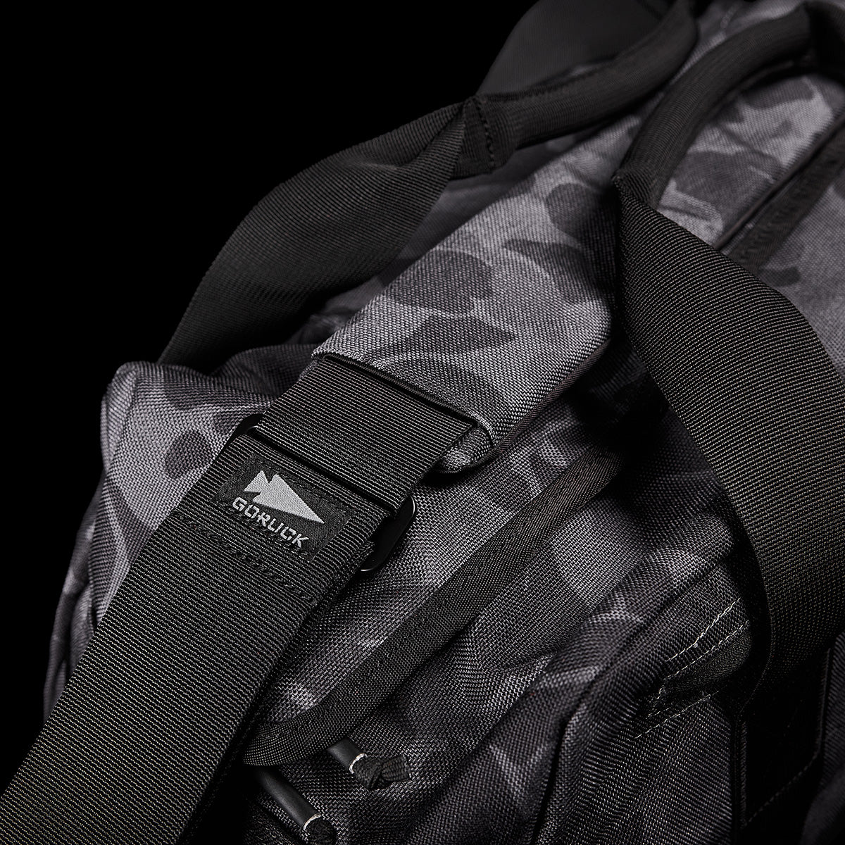 Close-up of the GORUCK Kit Bag, featuring a black and gray camouflage pattern reminiscent of a Special Forces Flight Bag. It has sturdy black straps, includes a robust buckle, and bears the GORUCK logo tag, all set against a solid black background.