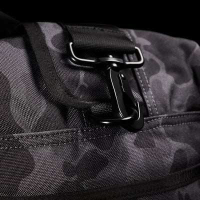 A close-up of a black and dark gray camouflage-patterned GORUCK Kit Bag (Includes Shoulder Strap) with a metal clasp and black strap. The fabric texture pops against the black background, evoking rugged functionality.