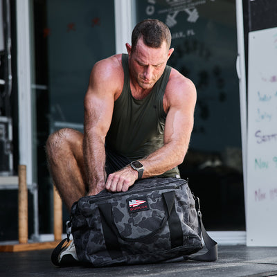 A man in a tank top crouches and unzips a GORUCK Kit Bag (Includes Shoulder Strap) outside a gym. He appears focused, with gym equipment and a chalkboard in the background, as if prepping for his next intense workout mission.