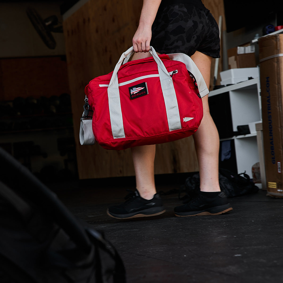 A person wearing black athletic shoes and shorts is holding a red and gray Kit Bag from GORUCK in a room with wooden shelving and cardboard boxes, suggesting they are either entering or leaving a fitness area.