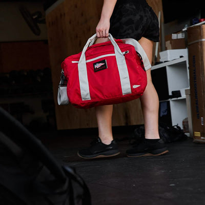 Carrying a Kit Bag (Includes Shoulder Strap) in bright red with white straps, a person paces through a room adorned with wooden and cardboard elements.