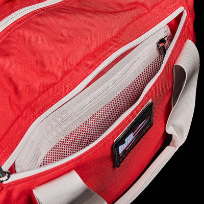 Close-up of a GORUCK Kit Bag (Includes Shoulder Strap) in red, resembling an Aviator’s Kit Bag, with a partially open zipper revealing an inner mesh pocket. The bag is styled with white handles and has a small black label on the front, enhancing its sleek design.