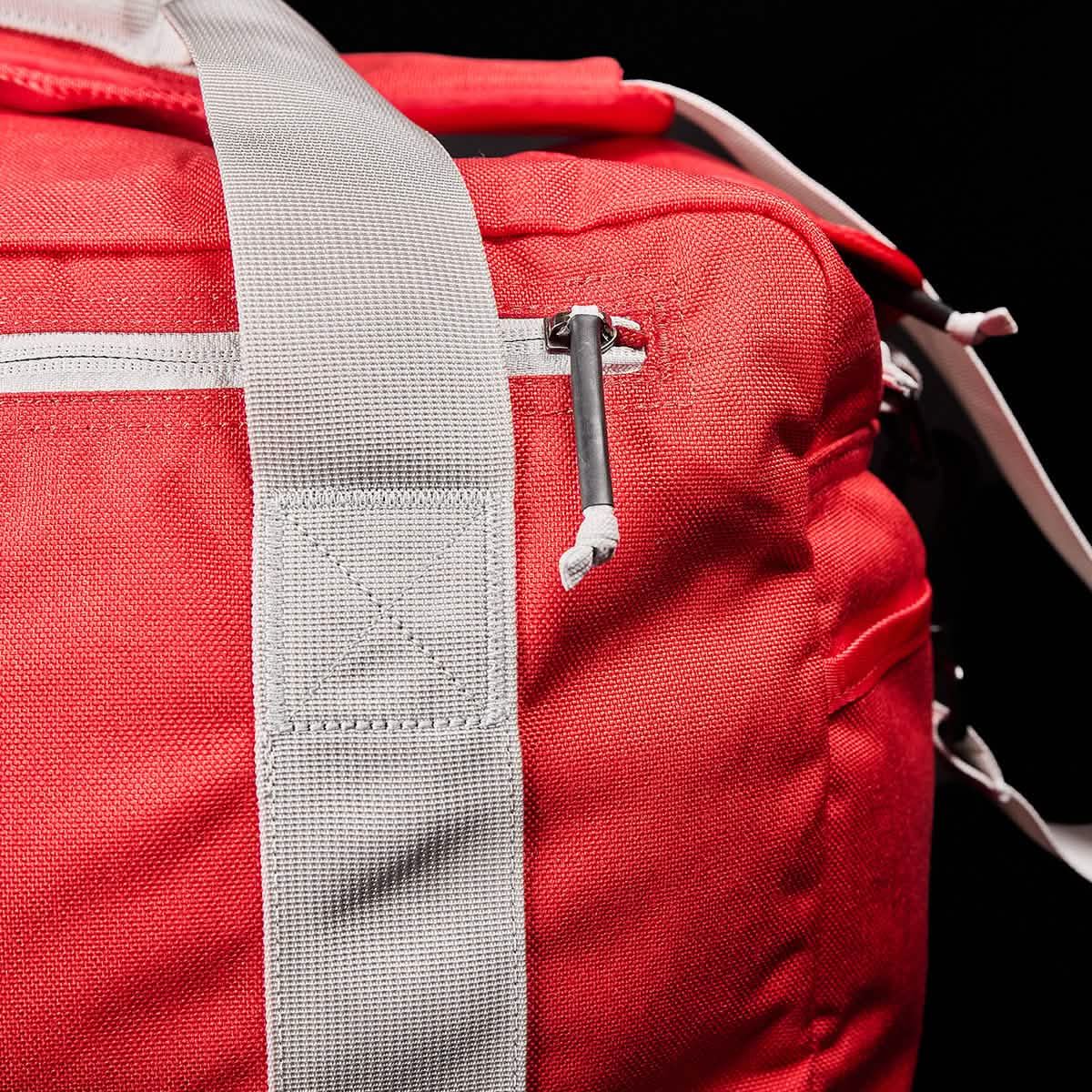 Close-up of a red Kit Bag from our collection, resembling an Aviator’s Kit Bag and featuring a white shoulder strap with a zippered pocket, set against a black background.