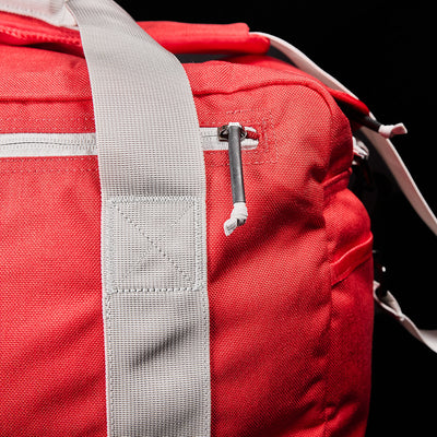 Close-up of a red GORUCK Kit Bag with white straps and a zipper. The fabric's texture is visible, and the zipper is equipped with a small black pull tab. The black background accentuates the vibrant color and distinctive details of this unique bag, complete with its shoulder strap.