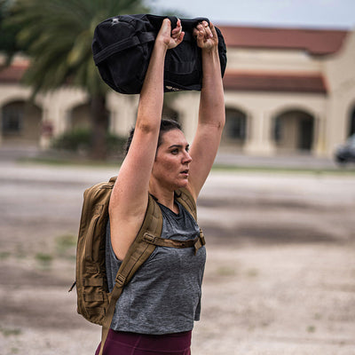 A woman in workout attire, sporting a Women’s USA Performance Tank - ToughMesh by GORUCK and carrying a backpack, lifts a black sandbag above her head while outdoors. The ToughMesh™ fabric ensures quick drying. In the background, a building with arches and palm trees enhances her focused and determined exercise routine.