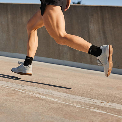 A woman jogging on a concrete surface wears black shorts and socks paired with the Women's Ballistic Trainers in Lunar Rock with Gum and Silver Reflective Spearhead by GORUCK. The image emphasizes the motion of her legs and muscular build, set against a concrete barrier under a clear sky. These trainers are ideal for lovers of functional fitness footwear.