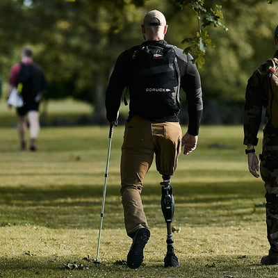 A person with a prosthetic leg walks through a grassy park with the aid of a walking stick, wearing GORUCK Mackall shoes in Black and Forged Iron for enhanced support. They have on a backpack and a cap. Nearby, someone in camouflage gear is partially visible, while trees and other walkers dot the background.