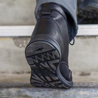 A close-up of the sole of a MACV-1 Traveler shoe by GORUCK highlights its All-Terrain Outsole's tread pattern as someone steps onto a concrete surface. The design on the sole features geometric shapes and lines, making it ideal for urban adventures.