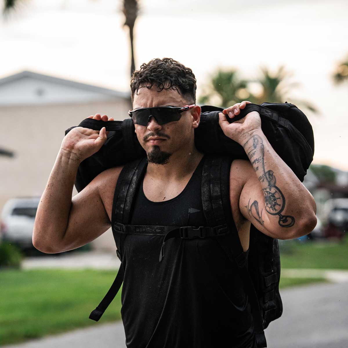 A man in a black Men's Performance Tank - ToughMesh by GORUCK and sunglasses carries a large backpack over his shoulders. He has tattoos on his arm and is walking outside on a sunny day, with blurred palm trees and houses in the background.