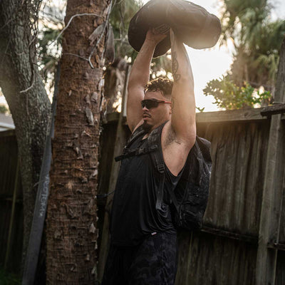 A person sporting sunglasses and the Men's Performance Tank - ToughMesh from GORUCK hoists a hefty bag above their head in an outdoor setting. With a backpack slung over their shoulders, they stand before a wooden fence and trees, sunlight filtering through the leaves, as the ToughMesh fabric ensures they stay cool and prepared for action.