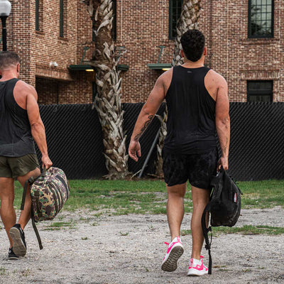 Two individuals dressed in athletic attire walk away from the camera, wearing Men's Performance Tanks made from ToughMesh fabric by GORUCK. They are carrying gym bags, with one sporting pink and white sneakers. The backdrop of a brick building and black fence enhances the scene's tough yet stylish vibe.