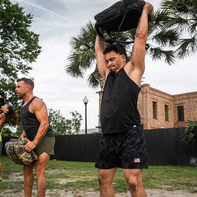 Two men enjoy exercising outdoors with one lifting a backpack over his head and the other holding a bag at waist height. Clad in GORUCK Men's Performance Tanks - ToughMesh and black athletic shorts, they revel in the fresh air surrounded by trees and a building in the background.