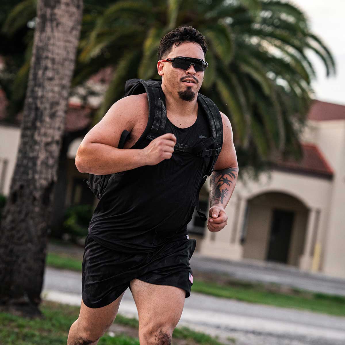 A person jogging with a backpack is wearing sunglasses, shorts, and the Men's Performance Tank - ToughMesh by GORUCK. The durable ToughMesh™ fabric of their gear whispers resilience as they pass palm trees and a building in the background.