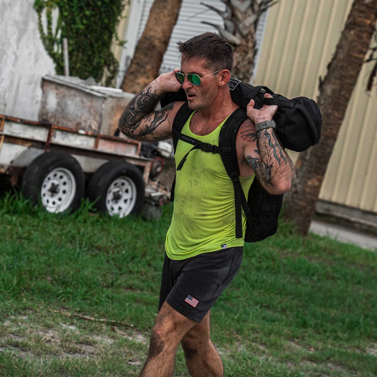 A man in a yellow GORUCK Men's Performance Tank made with ToughMesh™ fabric and black shorts carries a heavy black bag on his shoulders. Flaunting his tattoos and sunglasses, he exercises outdoors on grass near a trailer and palm trees, embracing the challenge.