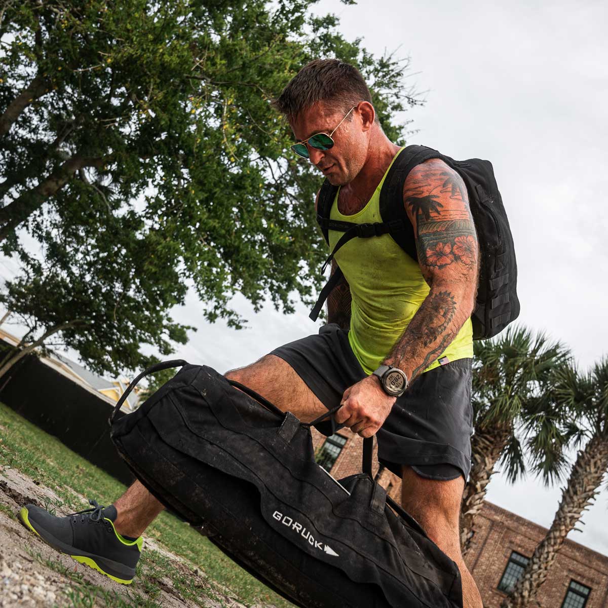 A man clad in a vibrant yellow Men's Performance Tank made from ToughMesh by GORUCK and sporting sunglasses is outdoors, hoisting a large black bag branded with GORUCK. Adorned with tattoos on his arms, he carries a backpack against the backdrop of trees and a building during his rigorous workout.