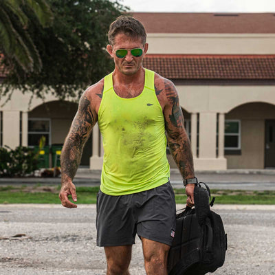 A man wearing a bright yellow Men's Performance Tank - ToughMesh by GORUCK and gray shorts walks outdoors, carrying a black bag. He has tattoos on his arms and wears sunglasses. The background shows a building with arches and a tree.