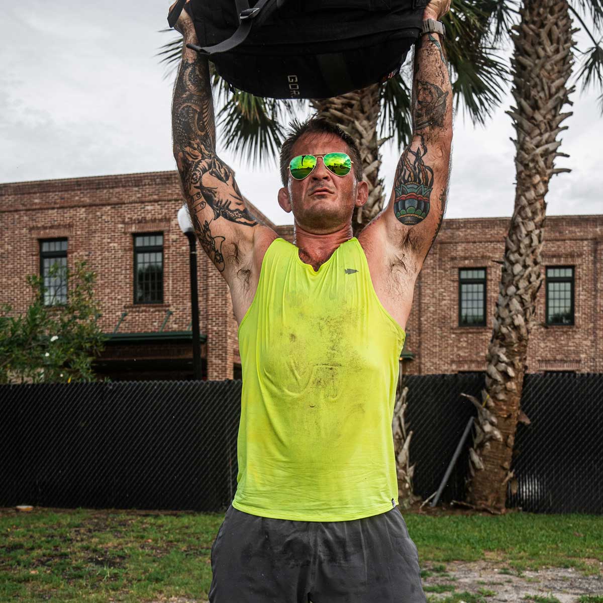 A man sporting a vibrant yellow GORUCK Men's Performance Tank - ToughMesh hoists a hefty sandbag overhead. His tattooed arms gleam under the sun, accented by reflective sunglasses. Standing amid brick buildings and palm trees, his attire embodies both grit and style, even as dust swirls around him.