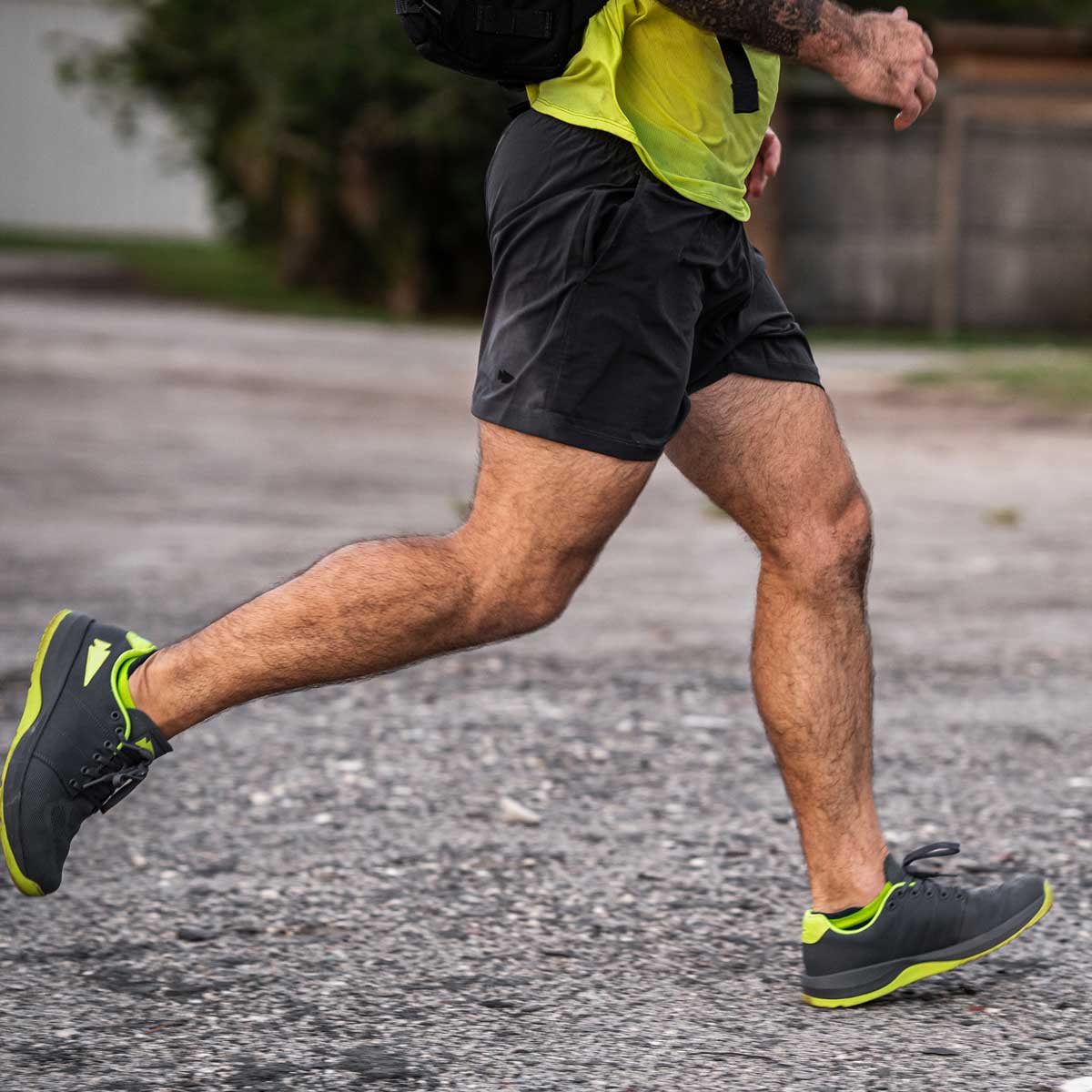 A person in a yellow shirt and GORUCK Men’s USA Training Shorts - ToughStretch is running on a gravel path. They are sporting gray and neon green running shoes. The background is slightly blurred, highlighting the freedom of movement provided by the ToughStretch fabric.