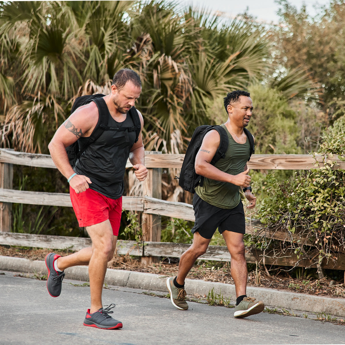 Two men jog on a paved path surrounded by greenery, each wearing athletic gear and backpacks. The first man sports a black tank top paired with red shorts, while the second is clad in a green tank top and black shorts. Their GORUCK Men's Ballistic Trainers in Ranger Green + White with Coyote Reflective Spearhead provide support for every stride, as tall grasses and a wooden fence line the background.