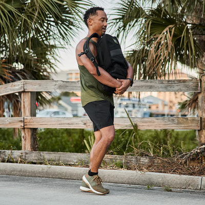 Wearing athletic apparel, a man stands outdoors on a sunny day, holding a backpack made from black CORDURA Ballistic Nylon. He's near a wooden fence with palm trees swaying in the background, epitomizing the essence of GORUCK's Men's Ballistic Trainers in Ranger Green and White with Coyote Reflective Spearhead—perfect for tropical or coastal adventures.