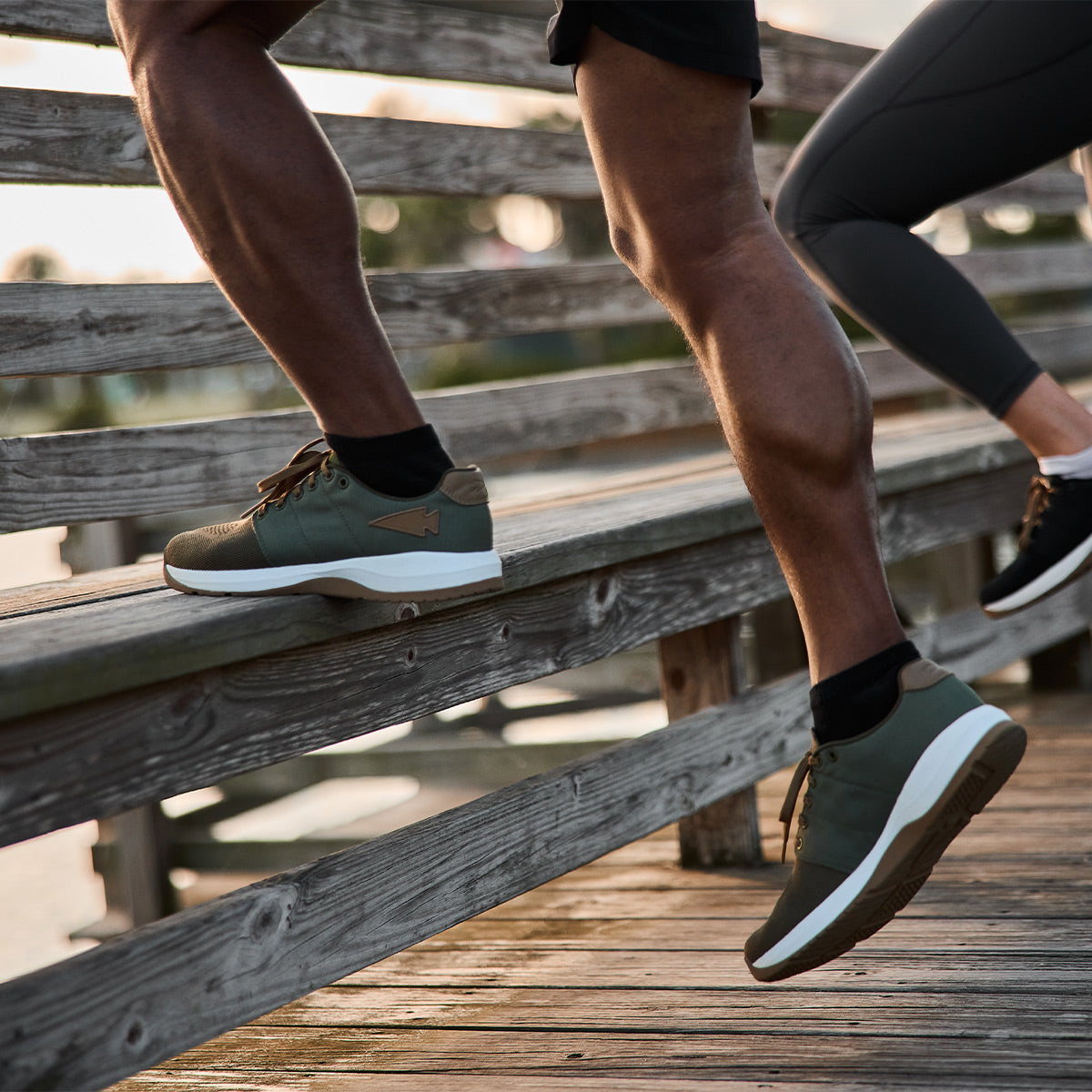 Two individuals dash up wooden steps in functional fitness footwear. One is wearing GORUCK's Men's Ballistic Trainers in Ranger Green with durable CORDURA Ballistic Nylon and white soles featuring a coyote reflective spearhead, while the other dons black sneakers equipped with 3X Support for stability. The scene takes place on an outdoor wooden structure or bridge.