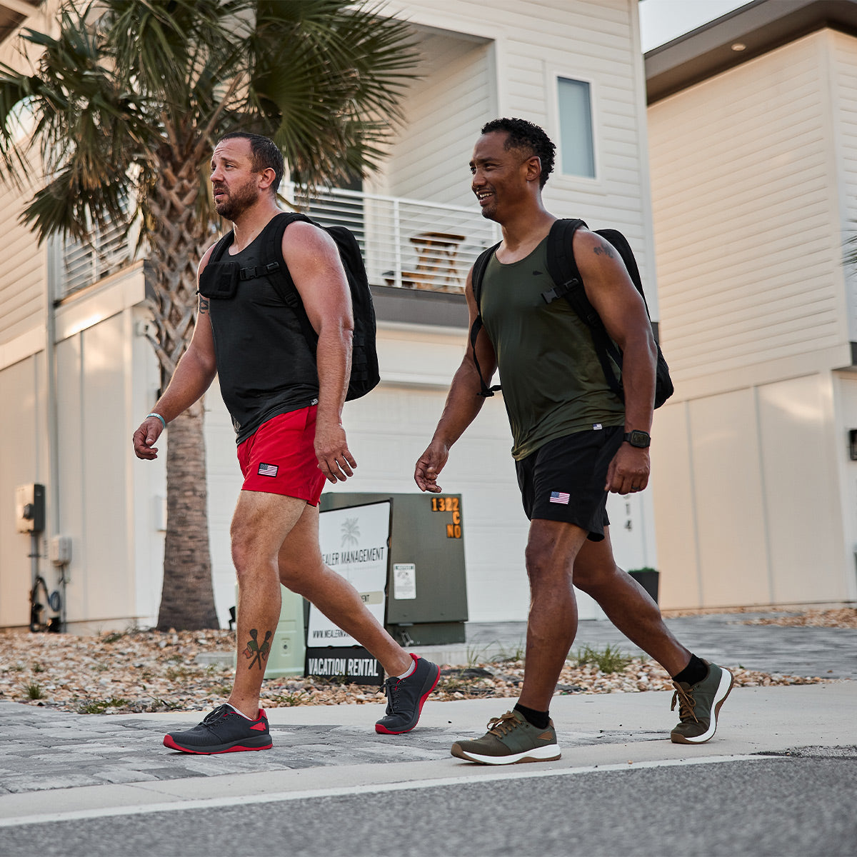 Two men walking on a sidewalk in front of beach rental homes. One wears a black tank top and red shorts, the other a green tank top and black shorts. Both carry GORUCK Men's Ballistic Trainers - Ranger Green + White W / Coyote Reflective Spearhead backpacks and appear to be enjoying a casual outdoor stroll.