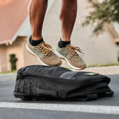 A person wearing the GORUCK Men's Ballistic Trainers in Ranger Green and White with Coyote Reflective Spearhead leaps over a black duffel bag on an outdoor surface. The background reveals part of a building, a tree, and a clear sky, with the durable CORDURA Ballistic Nylon ensuring their sneakers endure every dynamic move.