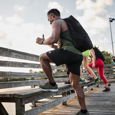 A person wearing a backpack, tank top, and shorts exercises outdoors on a wooden walkway, stepping onto a bench in their Men's Ballistic Trainers by GORUCK, featuring Ranger Green and White with Coyote Reflective Spearhead accents. Two others are in the background. The sky is partly cloudy on this sunny day.