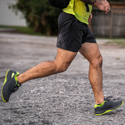 A person in a neon green shirt and black shorts is running on a paved path, showcasing their Men's Ballistic Trainers - Wolf Grey + Acid Lime W / Acid Lime Reflective Spearhead from GORUCK. Their focus highlights the 3X Support features, while the blurred background captures their swift motion.