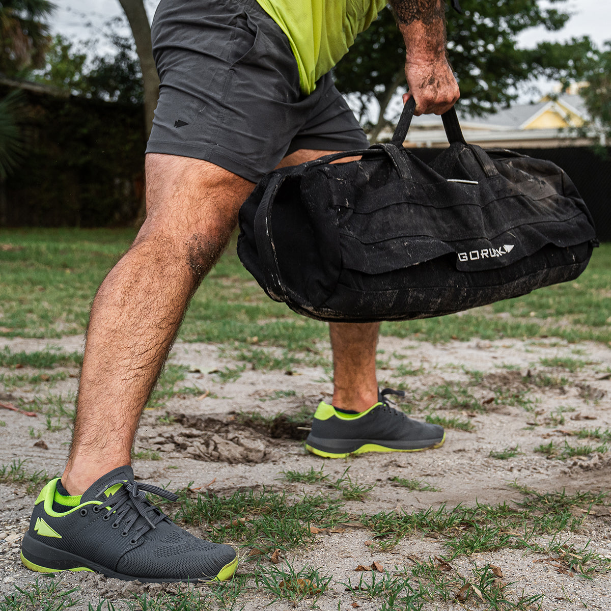 A person in wolf grey shorts, complemented by Men's Ballistic Trainers in Wolf Grey and Acid Lime with the distinctive reflective spearhead design, is lifting a robust black duffel bag crafted from CORDURA Ballistic Nylon, proudly displaying the GORUCK brand. The scene unfolds outdoors amidst grass, dirt, and a backdrop of trees.