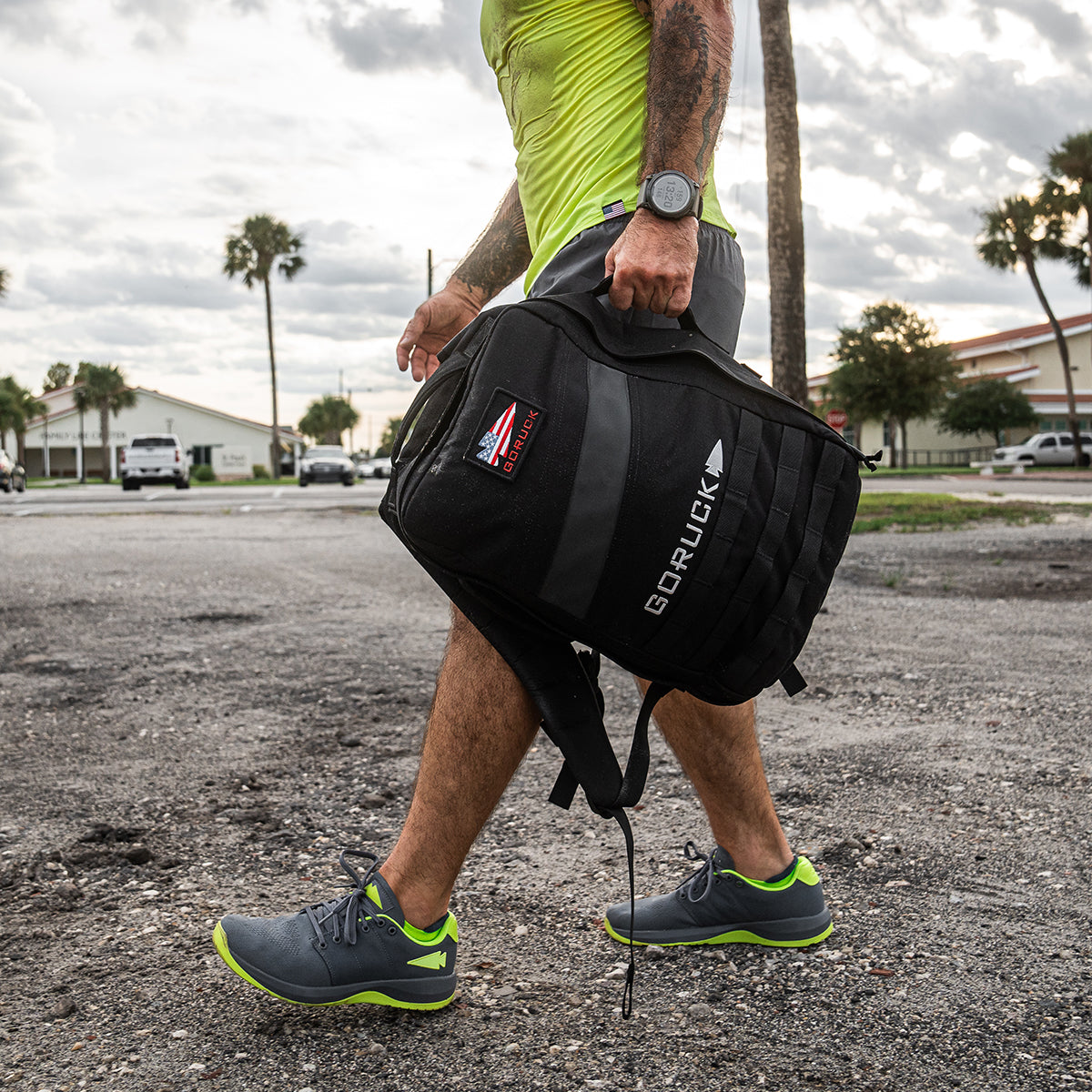 A person wearing GORUCK's Men's Ballistic Trainers in Wolf Grey and Acid Lime, featuring the Acid Lime Reflective Spearhead design, strolls on a gravel path with a backpack crafted from durable CORDURA Ballistic Nylon. The sky is overcast, and trees and buildings are visible in the background.