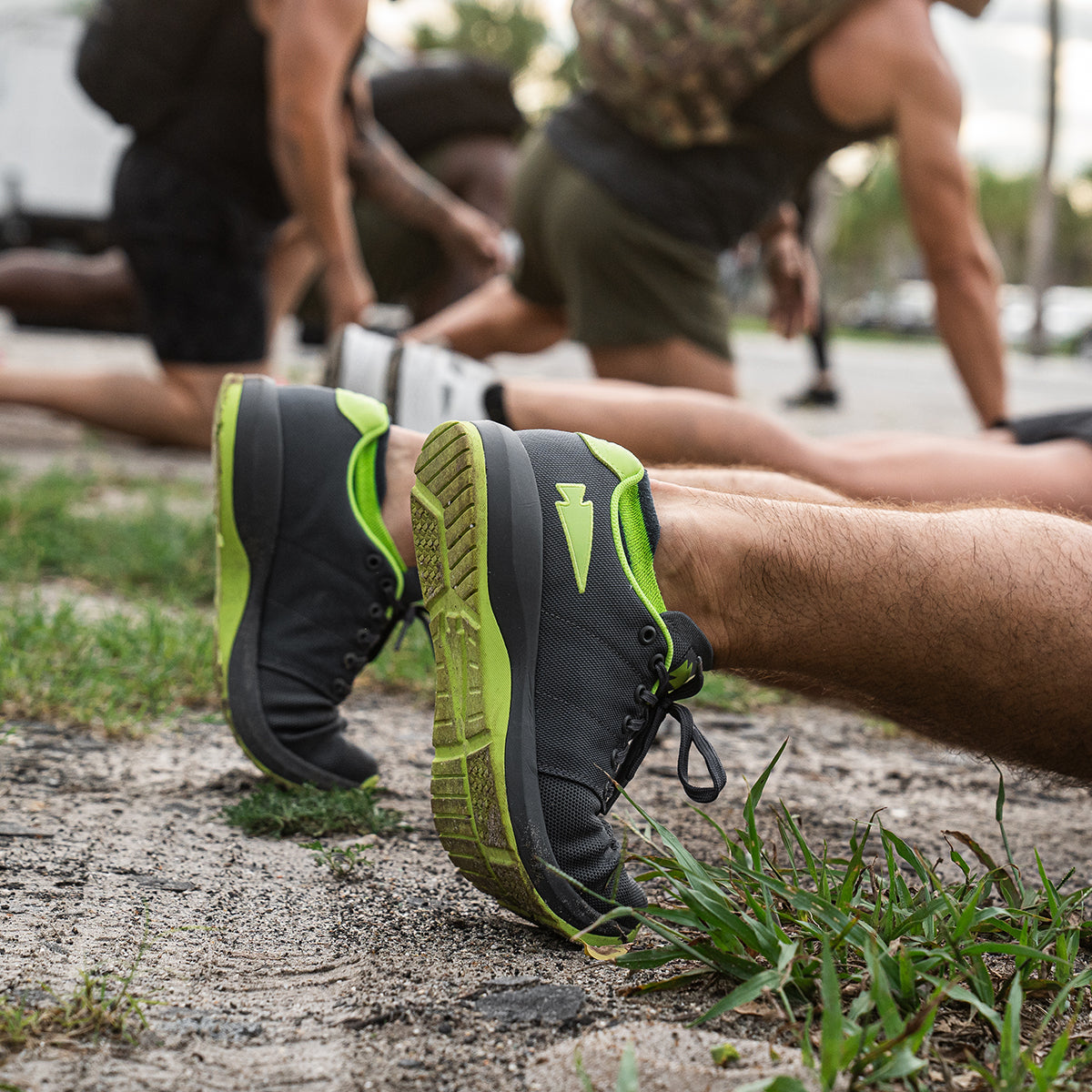 A group of people exercising outdoors showcases an individual wearing the Men's Ballistic Trainers in Wolf Grey and Acid Lime by GORUCK, designed for functional fitness. As they stretch on the ground, other participants blur in the background while grass frames the scene.