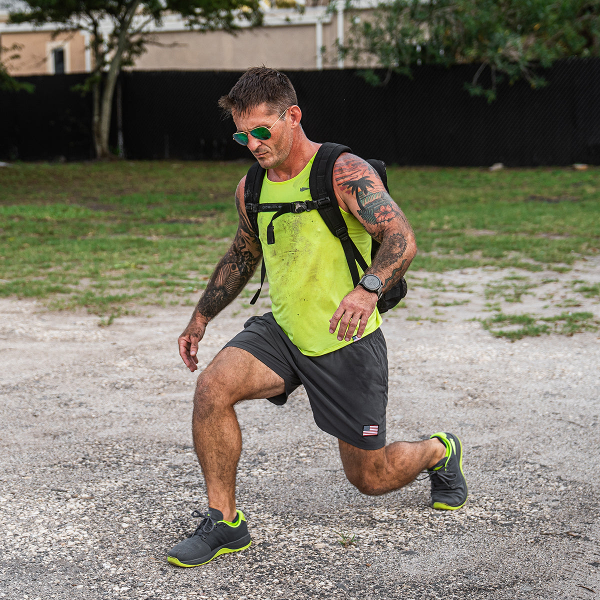 A man wearing a vivid green sleeveless shirt and gray shorts exercises outside, sporting GORUCK's Men's Ballistic Trainers in Wolf Grey and Acid Lime with Acid Lime Reflective Spearhead. He carries a CORDURA Ballistic Nylon backpack and dons sunglasses as he performs lunges on a gravel path, encircled by trees with a black fence in the background.